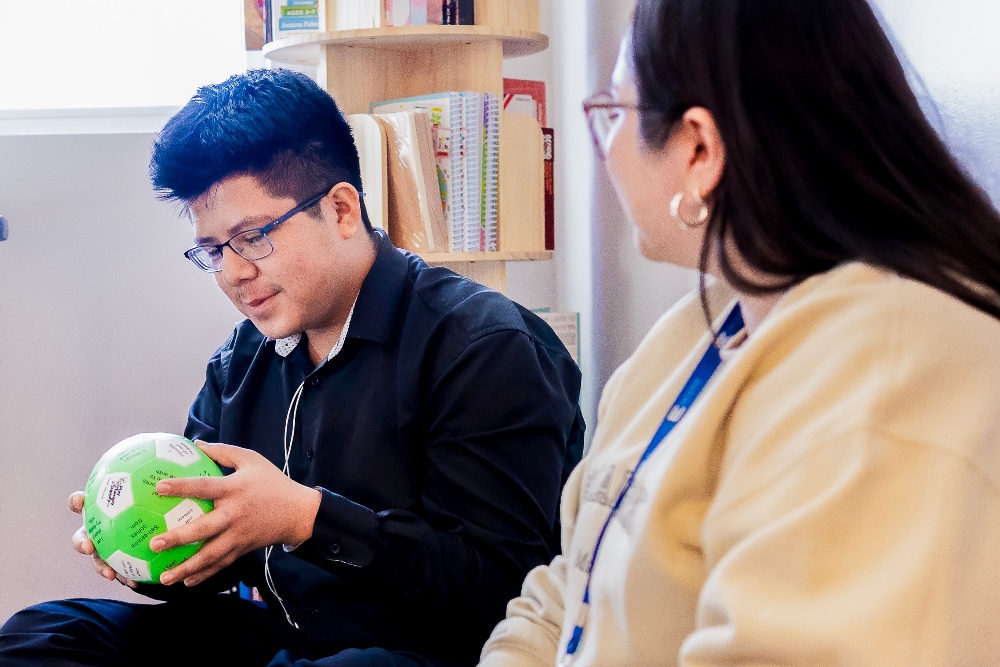 A male Empower College Prep high school student sitting next to his female teacher, looks down at a green and white ball printed with affirmations that he’s holding.